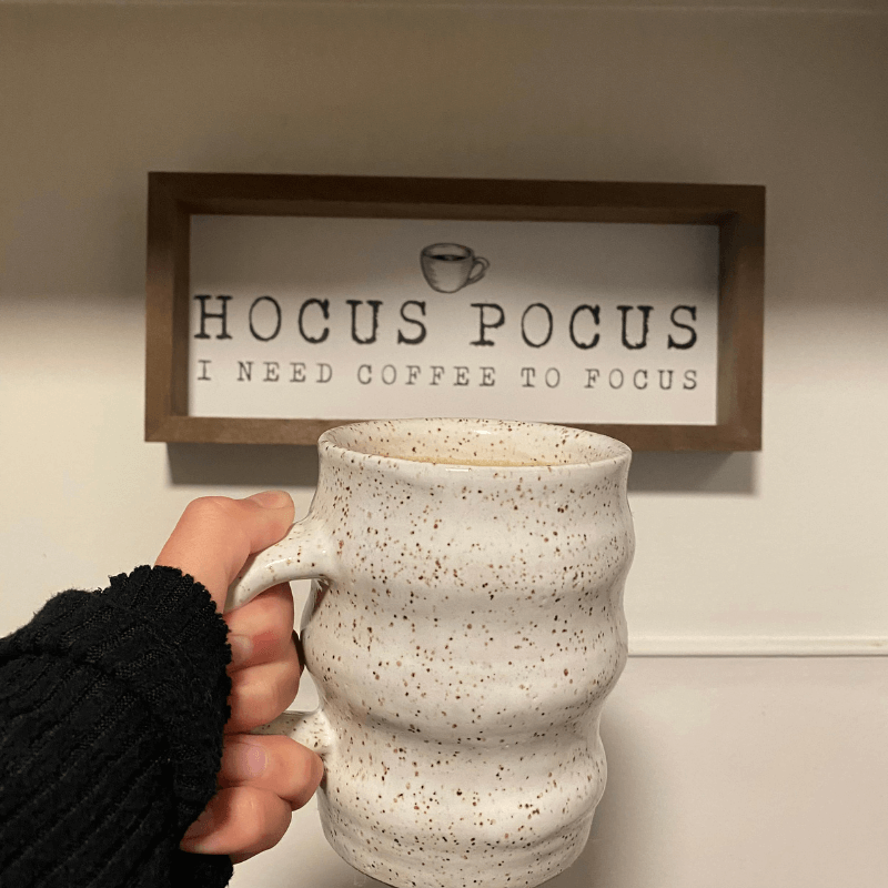 Favorite Things About Fall - Hand Holding a Mug in Front of a Sign Saying Hocus Pocus I Need Coffee to Focus
